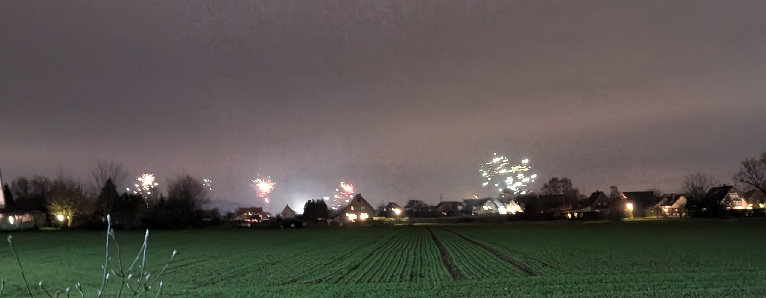 Hinter einem grünen Acker sieht man die Silhouette von Häusern und Bäumen. Einige Silvesterraketen steigen in den grauen, leicht nebeligen Himmel.